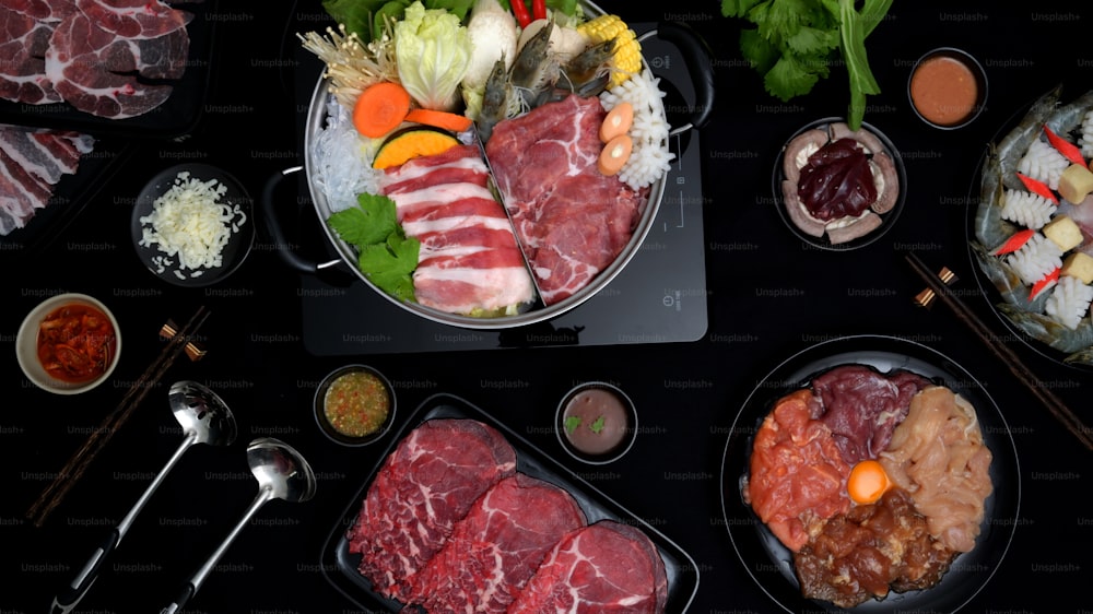 Vista superior de Shabu-Shabu em panela quente, carne fresca fatiada, frutos do mar, legumes e molho de mergulho com fundo preto, cozinha japonesa