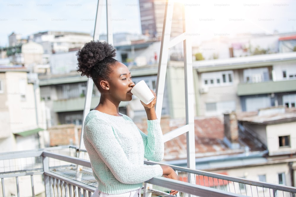 African American Woman drinking coffee on her balcony with a city view in the sun, outdoor in sunlight light, enjoying her morning coffee.