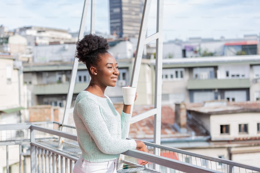 Retrato de uma mulher negra afro-americana feliz pensando e olhando para o lado com café nas férias com um resort ou hotel com vista da cidade ao fundo