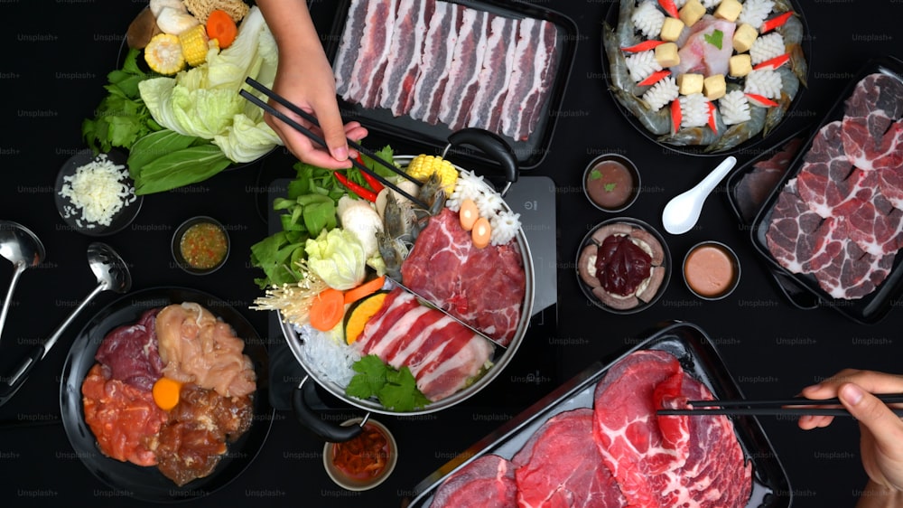 신선한 얇게 썬 고기, 바다 음식, 검은 배경의 야채, 일본 요리와 함께 냄비에서 샤브샤브를 먹는 사람들의 상위 뷰