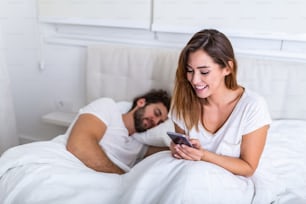 男性がベッドで寝ている間、携帯電話を見ている女性。若い美しい女性は、寝室のベッドで寝ている間、携帯電話でプライベートに話しています
