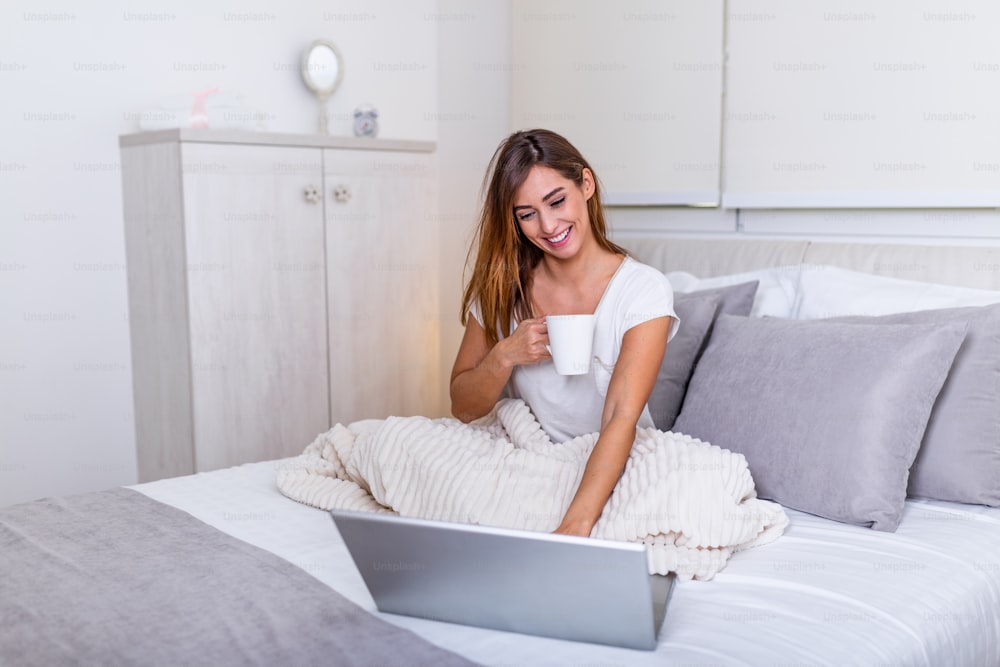 Hermosa joven bebiendo café en casa en su cama con una camiseta acogedora mientras trabaja en su computadora portátil. Revisar el correo electrónico por la mañana, concepto independiente