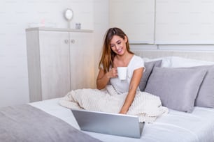 Beautiful young woman drinking coffee at home in her bed wearing a cozy t shirt while working on her laptop. Checking email in the morning, freelance concept