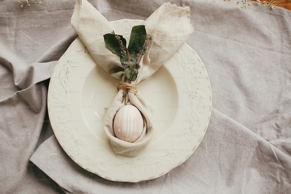 Stylish Easter brunch table setting with egg in easter bunny napkin. Modern natural dyed pink egg on napkin with bunny ears, lavender flowers on vintage plate. Easter table decorations
