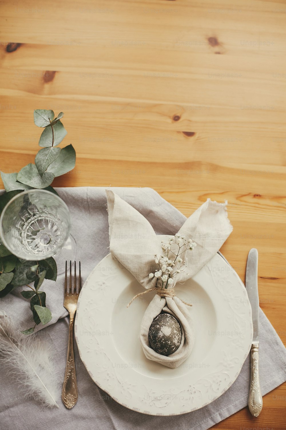 Easter table decorations. Modern natural dyed gray egg on napkin with bunny ears, flowers on vintage plate and cutlery. Stylish Easter brunch table setting with egg in easter bunny napkin.