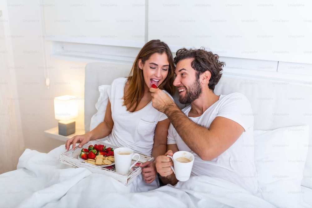 Verliebtes Paar frühstückt im Bett. Junges kaukasisches Paar beim romantischen Frühstück im Bett. Weiblich und männlich, zwei Tassen Kaffee, Obst und bunte Kekse.