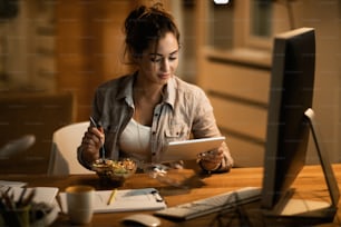 Smiling woman surfing the net on digital table while eating salad in the evening at home.