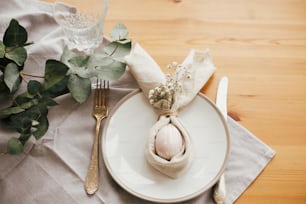 Stylish Easter brunch table setting with egg in easter bunny napkin on  table. Modern natural dyed pink easter egg on napkin with bunny ears, flowers on plate and cutlery. Easter table decorations