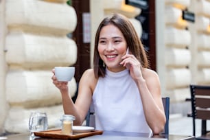 Mujer asiática sonriente bebiendo café y usando su teléfono móvil. Mujer satisfecha disfrutando de una taza de café. Retrato de cerca de una hermosa muchacha bebiendo café de una taza blanca en la cafetería