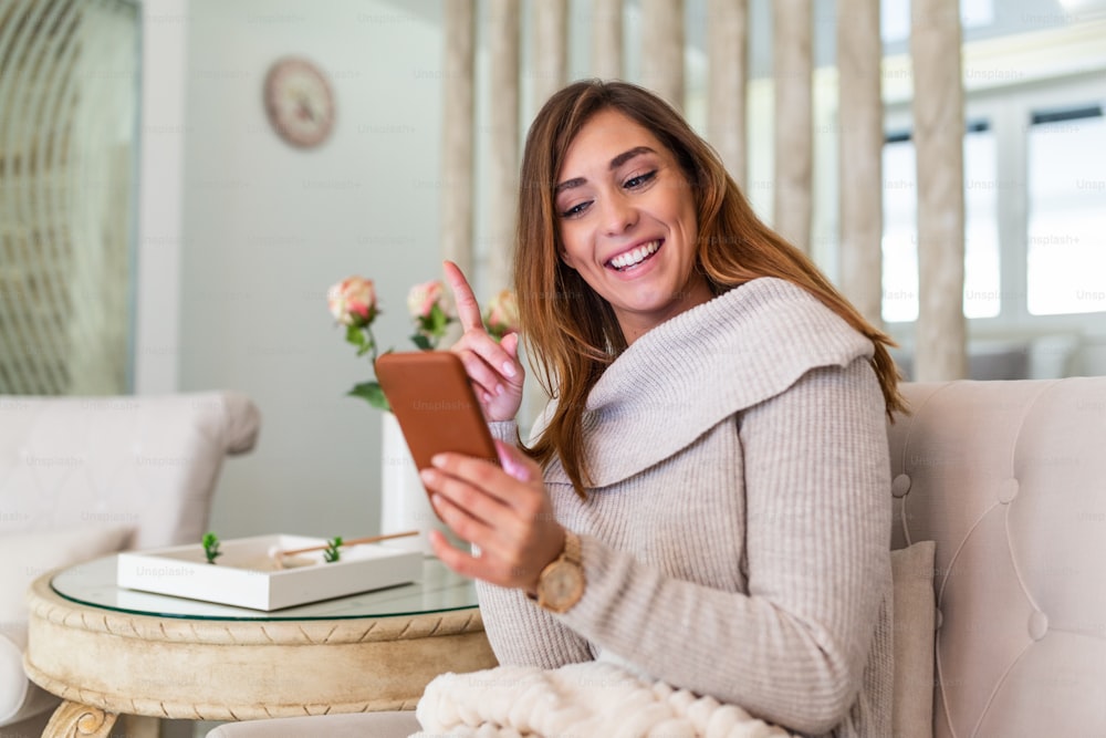 Portrait of beautiful young woman using her mobile phone at home. Lovely message from him. Beautiful young smiling woman looking at her smart phone while sitting in a big comfortable chair at home