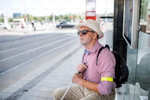 A senior blind man with white cane waiting for public transport in city.