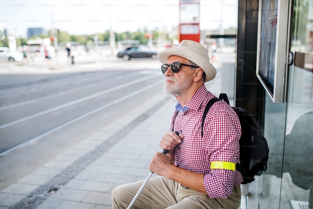 A senior blind man with white cane waiting for public transport in city.