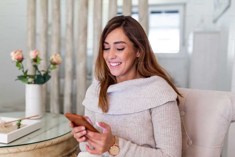 Lovely message from him. Attractive young woman looking at her smart phone and smiling while sitting on the sofa at home. Beautiful woman texting on her mobile phone at home