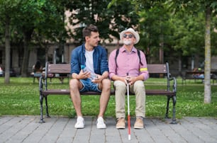 A young man and blind senior with white cane sitting on bench in park in city, talking.