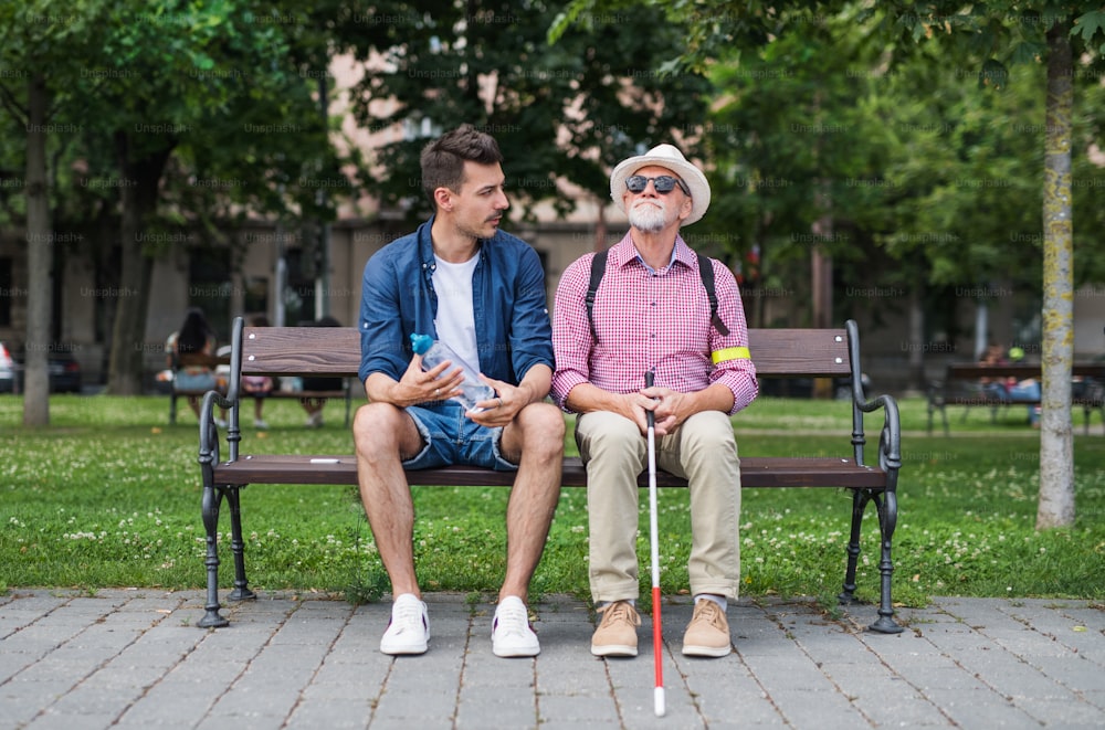 A young man and blind senior with white cane sitting on bench in park in city, talking.