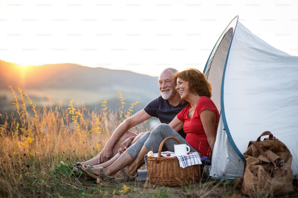 A senior tourist couple with picnic basket sitting in nature at sunset, resting.