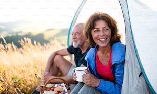 A senior tourist couple with picnic basket sitting in nature at sunset, resting.