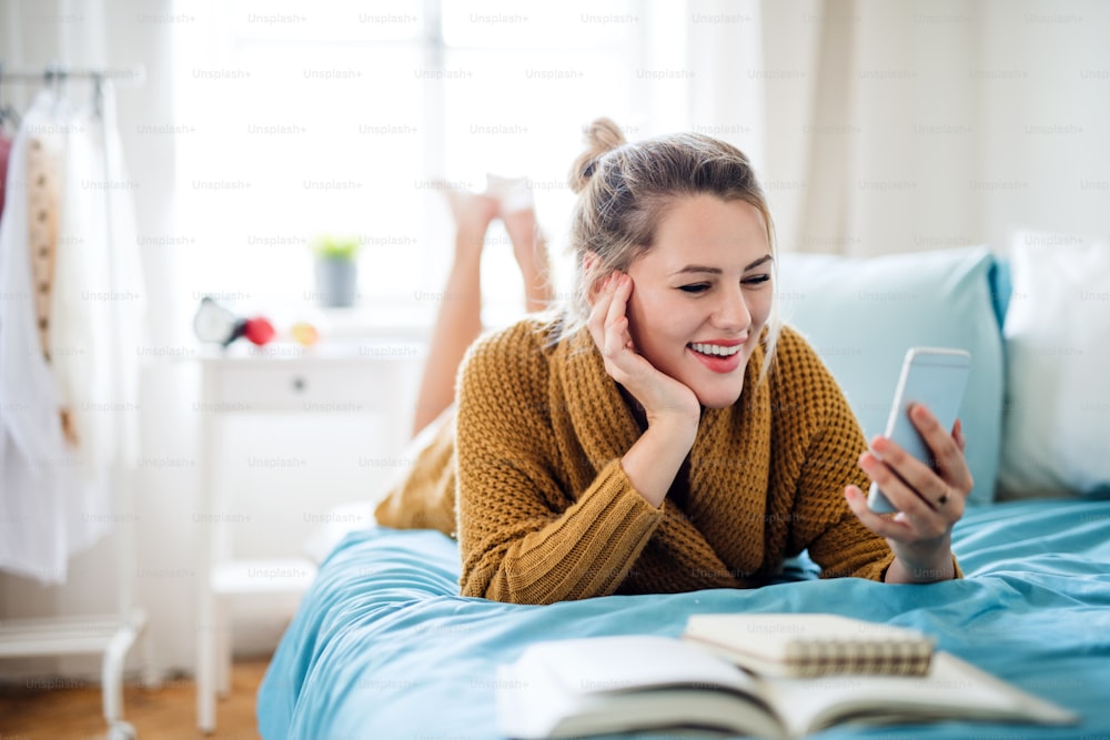 스마트폰을 들고 있는 행복한 젊은 여성이 집 실내 침대에 누워 휴식을 취하고 있다.