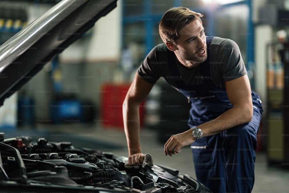 Young auto mechanic looking away while repairing car engine in auto repair workshop.
