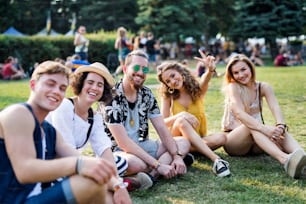 Group of cheerful young friends sitting on ground at summer festival, looking at camera.