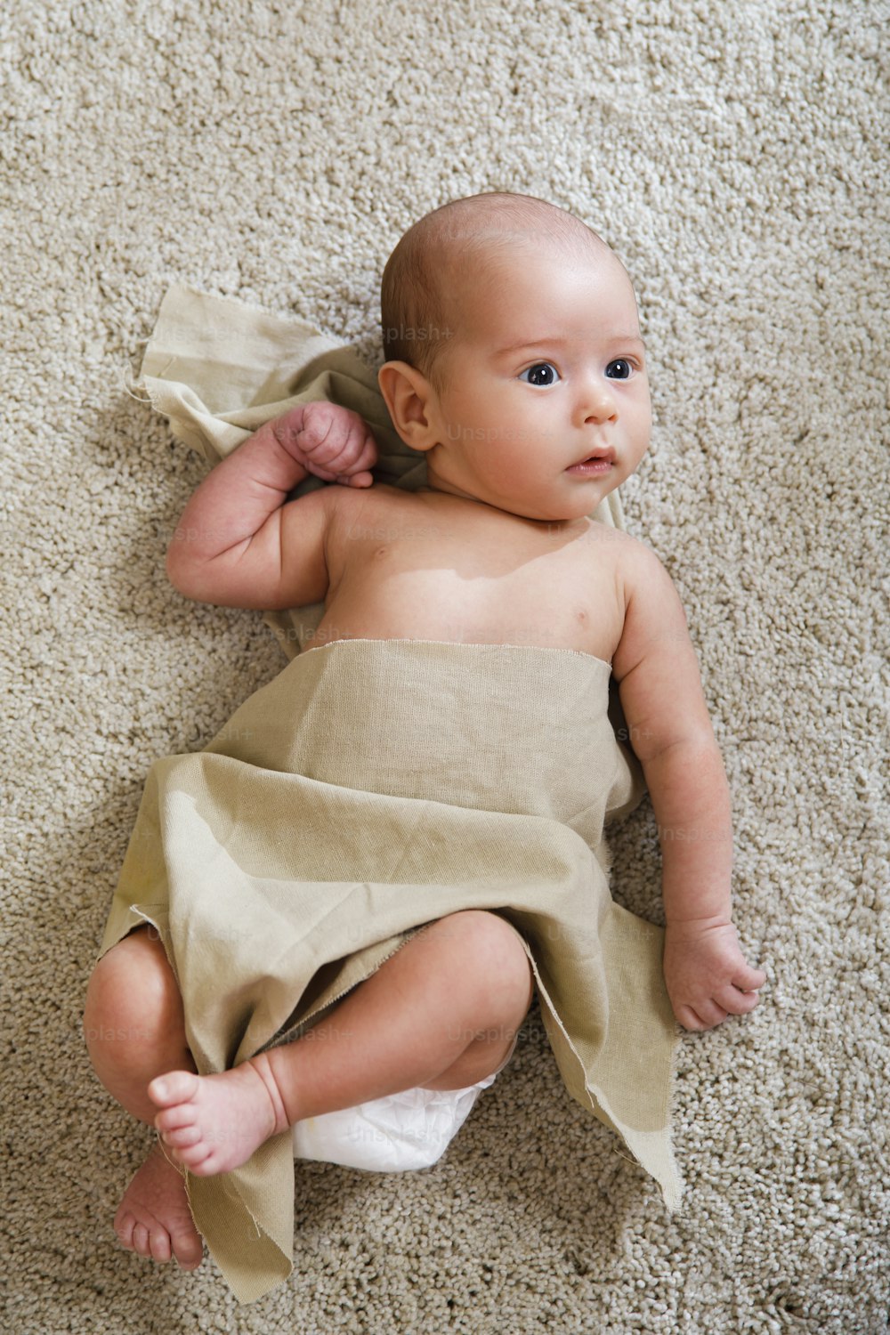 Cute little baby covered with piece of linen fabric is lying on the soft carpet