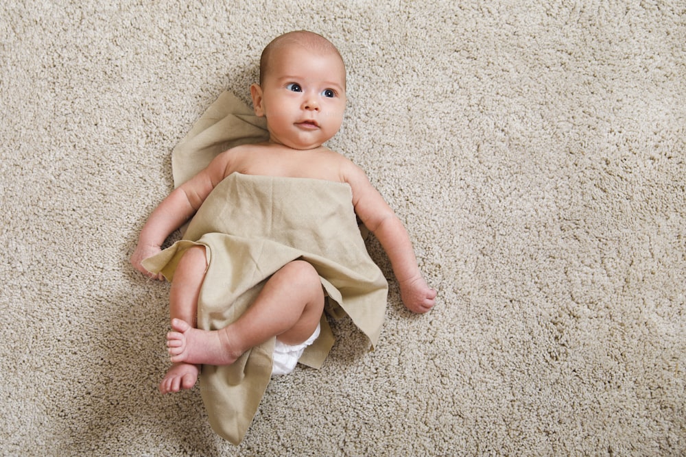 Cute little baby covered with piece of linen fabric is lying on the soft carpet
