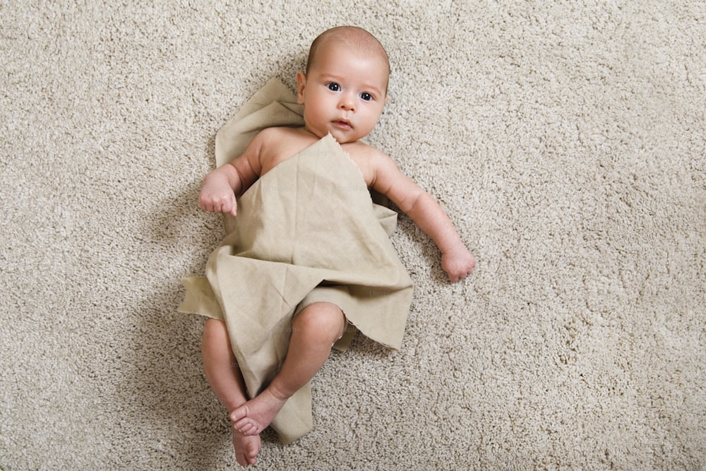 Cute little baby covered with piece of linen fabric is lying on the soft carpet