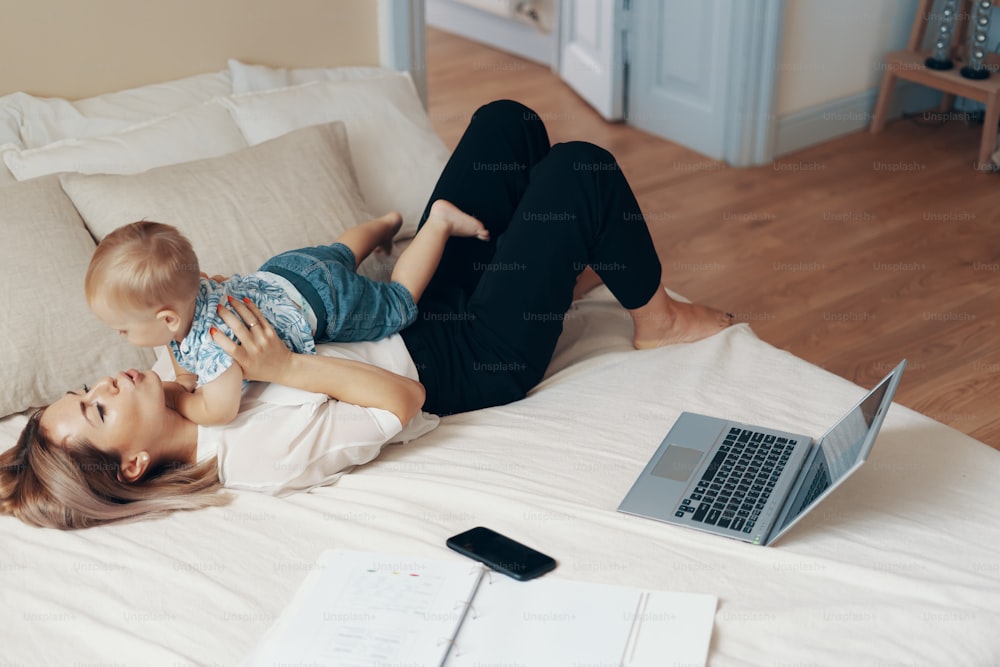 Young mother with her child working in bedroom at home. Business mom takes a break. Multi-tasking, freelance and motherhood concept