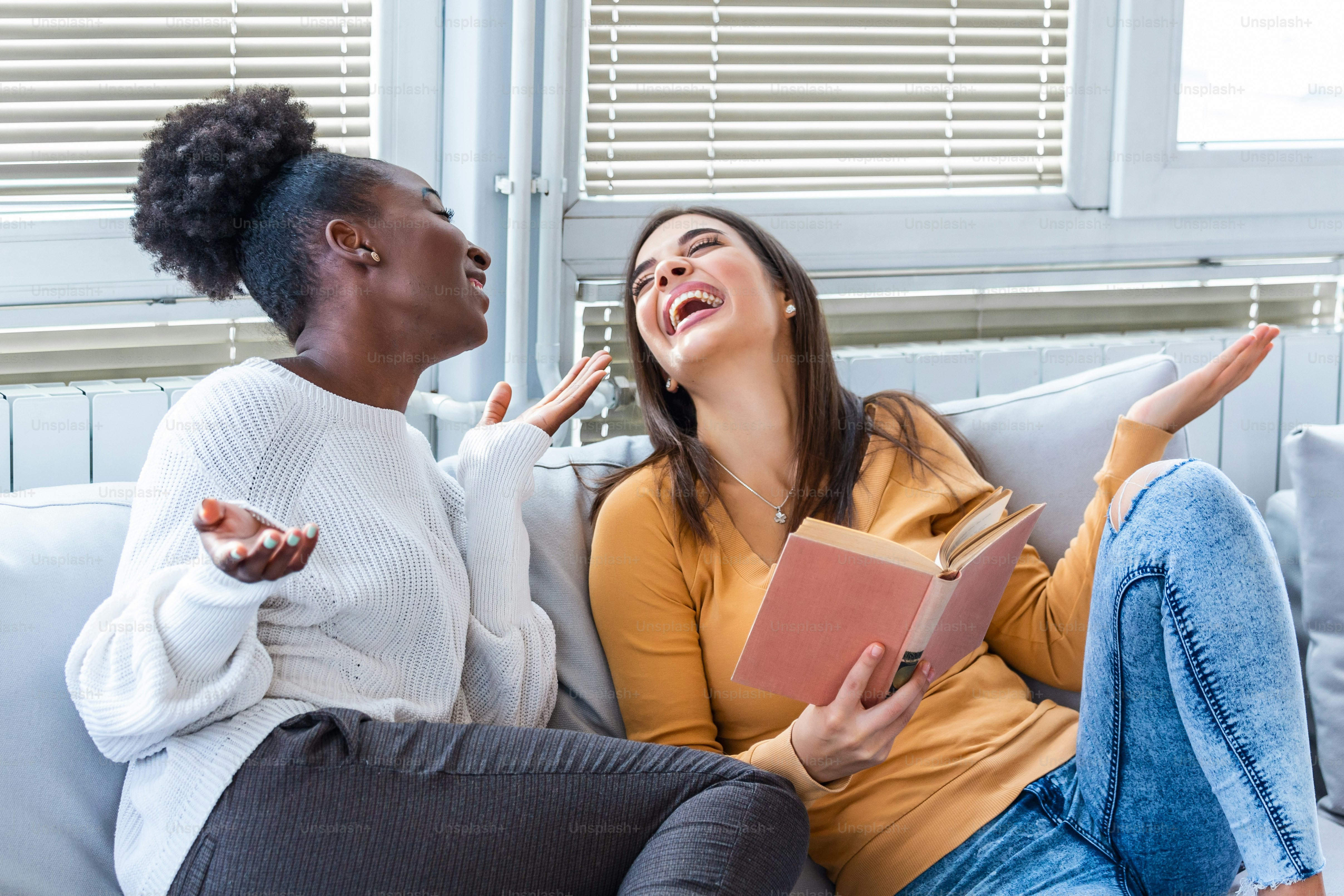 Two women laughing about funny stories from books at home. Friendship and care, happy girl friends. African American and caucasian best friends having fun at home, laughing and reading a funny book.