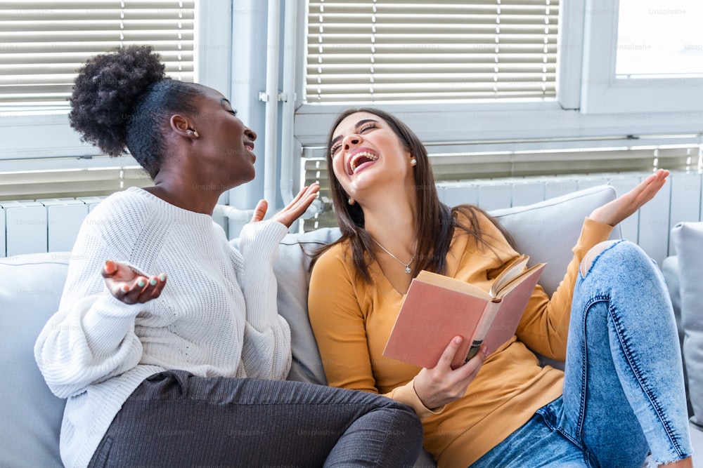 Two women laughing about funny stories from books at home. Friendship and care, happy girl friends. African American and caucasian best friends having fun at home, laughing and reading a funny book.
