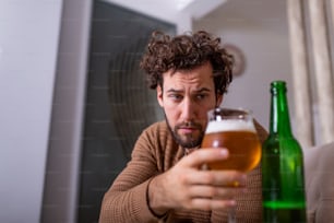 Nach der Arbeit sitzt ein depressiver Typ auf dem Sofa und trinkt ein kaltes Bier. Hand hält ein Glas, Mann trinkt allein nach Hause. Alkoholismus, Alkoholabhängigkeit und Menschenkonzept