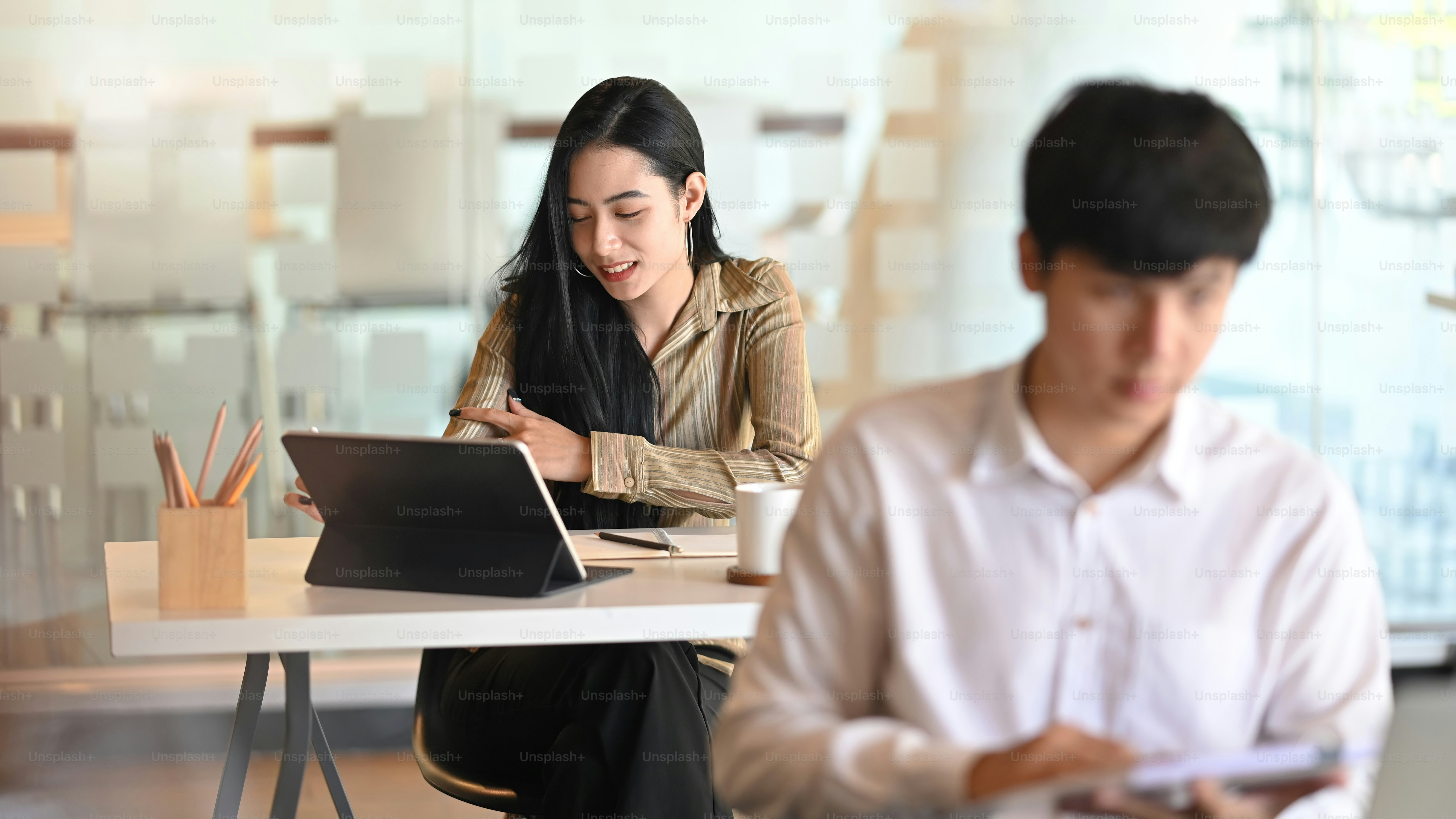 Young professional female designer working on her project while tablet with co-worker in the background.