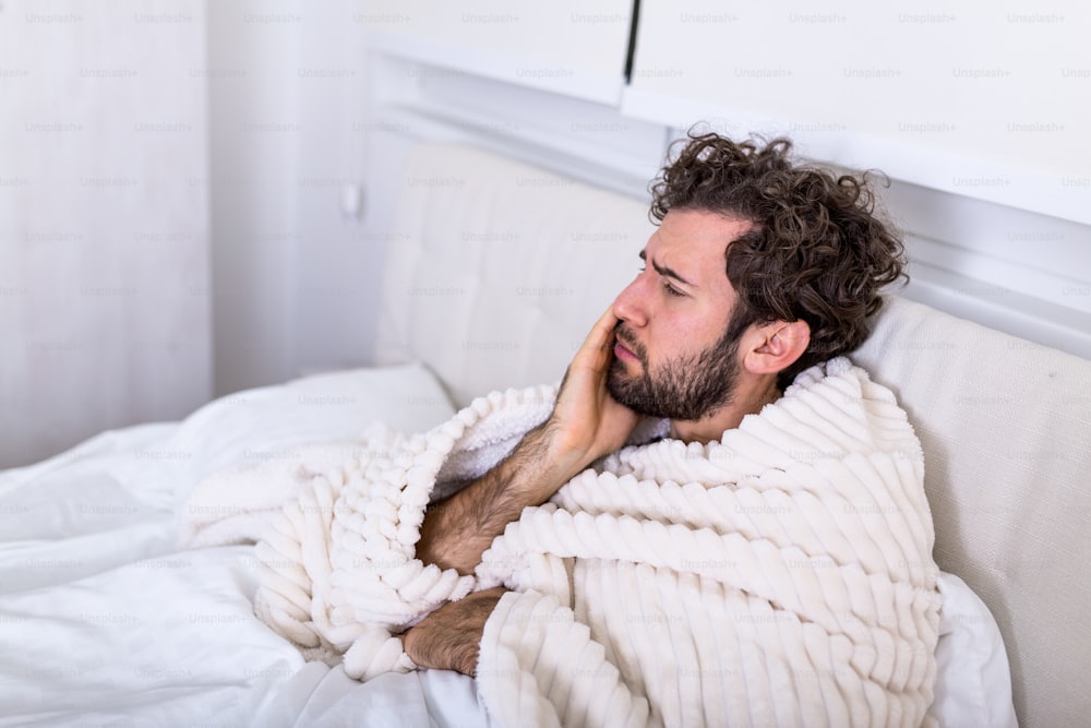 Tooth ache concept. Indoor shot of young male feeling pain, holding his cheek with hand, suffering from bad toothache, laying in bed with painful expression