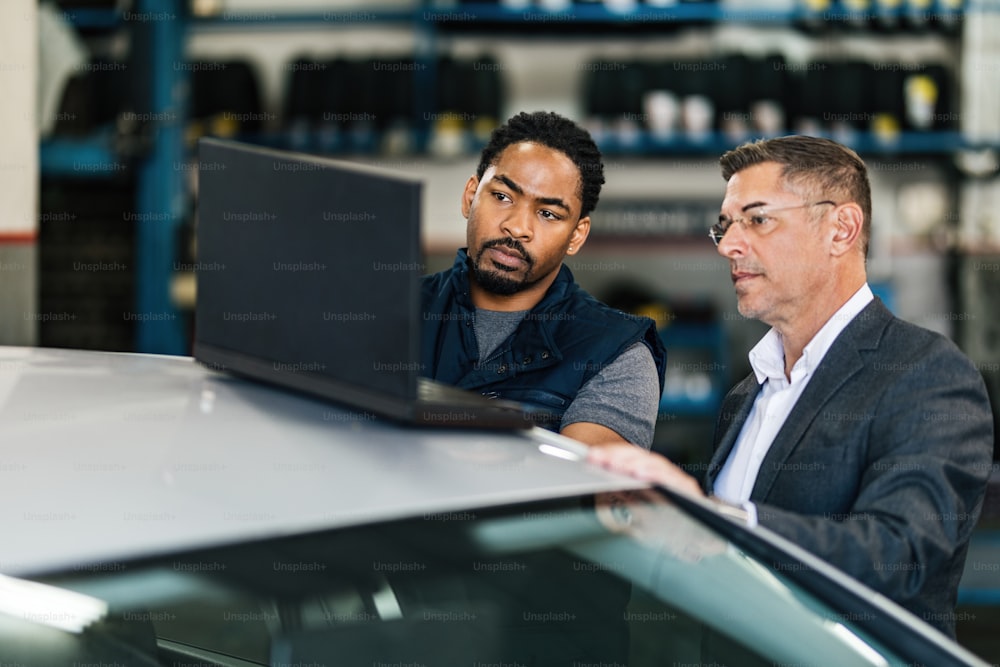 Young black auto mechanic and his customer cooperating while using computer in repair workshop.