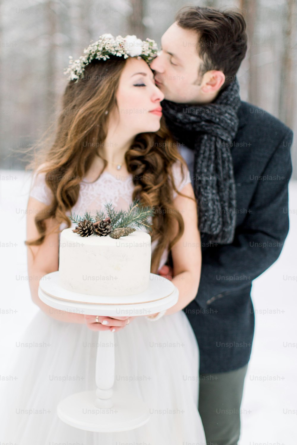 Beautiful wedding couple posing with decorated cake in hands standing outdoors in forest, groom kissing her bride. Winter snowy forest background.