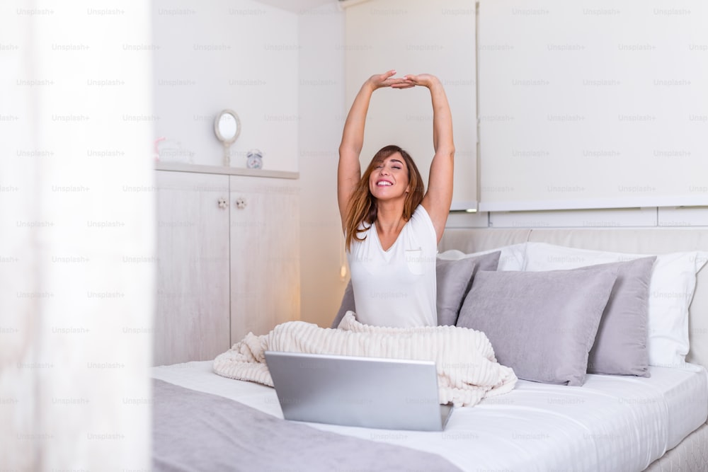 Woman stretching in bed after wake up in morning ready to work from home. Portrait of a young smiling woman sitting with laptop on bed. Freelance working from home concept