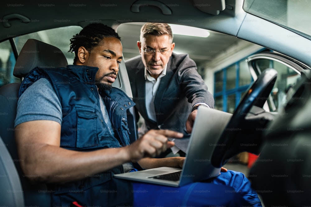 Young black mechanic and male customer using computer and pointing on something on the screen in auto repair shop.
