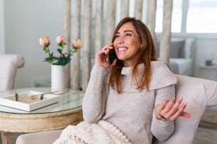 Happy young woman caller talking on her mobile phone at home, cheerful teen girl enjoys pleasant mobile conversation, smiling millennial female holding cell phone making call in living room