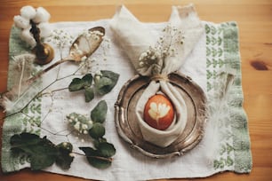 Stylish Easter brunch table setting with egg in easter bunny napkin. Modern natural dyed egg on napkin with bunny ears, flowers on vintage plate. Rustic Easter table decorations