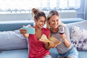 Friends eating pizza and smiling for selfie. They are sharing pizza and making selfie photo on mobile smart phone. They are having party at home, eating pizza and having fun.