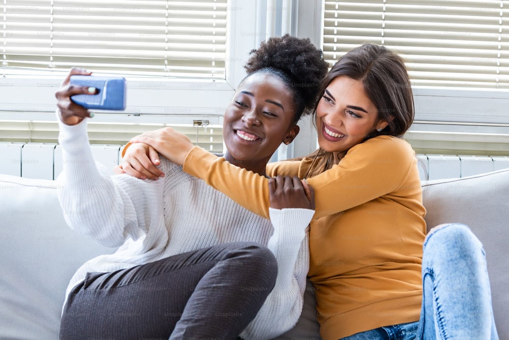 Laughing woman friends hugging each other on sofa while taking selfie photo on smart phone. Lovable caucasian and African American girls expressing positive emotions to camera.