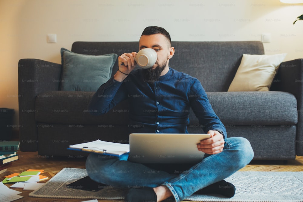 Man drinking coffee in the morning sitting on the floor at home while working at his laptop. Handsome male freelancing at home.