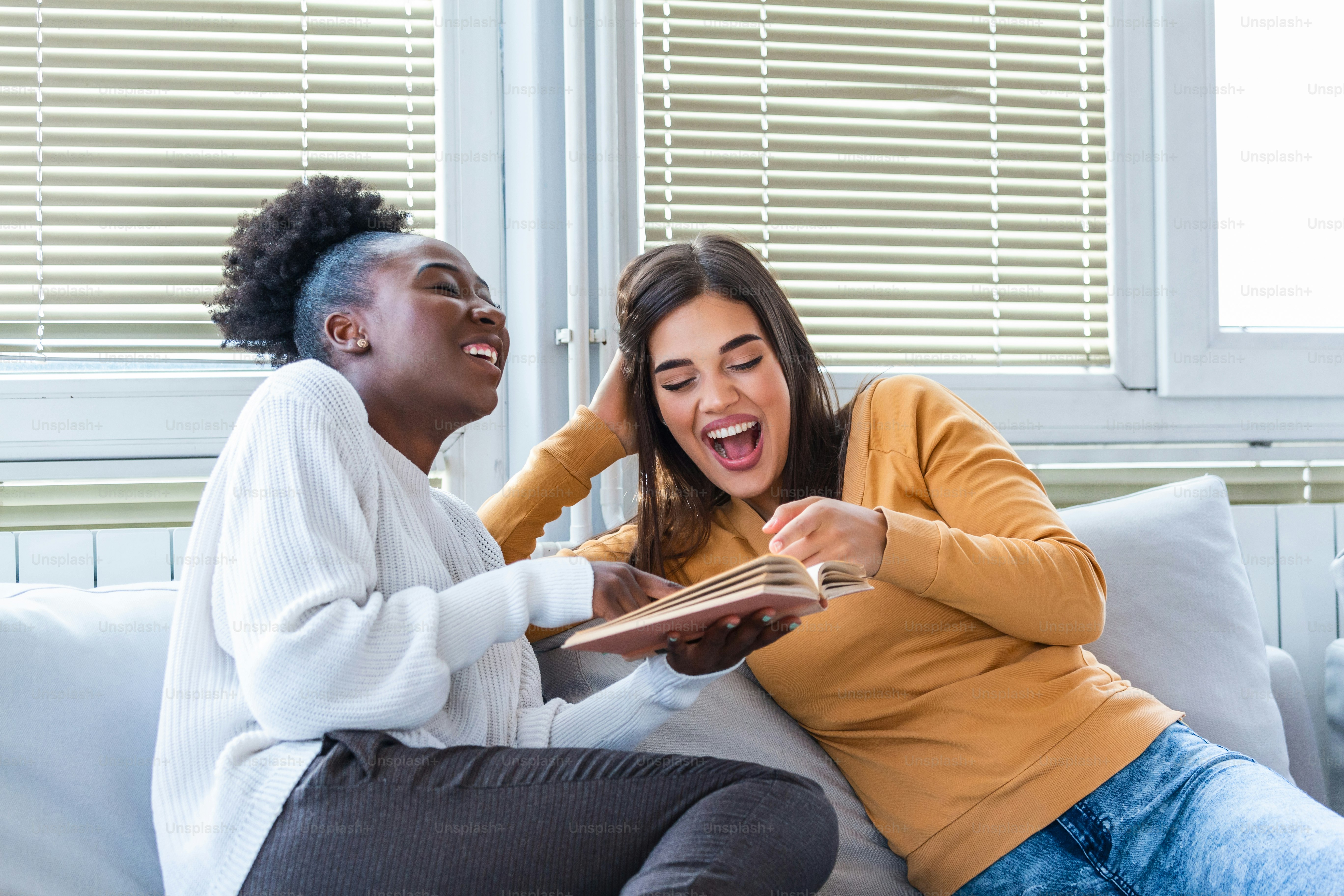 Girls having fun at home, laughing and reading a funny book. Different race friends at home reading a book and enjoying their time together