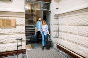 Pretty woman and handsome man holding hands are leaving a hotel with their suitcases.