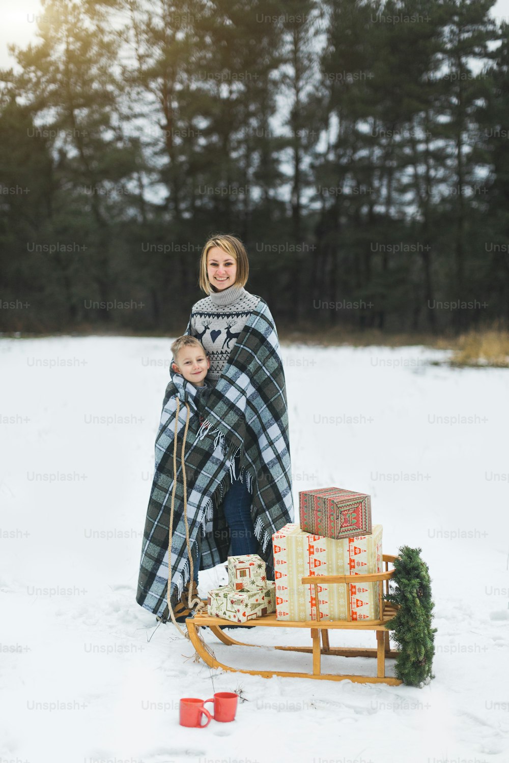 Hübsche junge Mutter mit ihrem Sohn, der mit kariertem Plaid bedeckt ist, über dem hinteren Teil des Winterwaldes stehend, geschmückten Holzschlitten mit Weihnachtskranz zieht und Geschenke darauf präsentiert.