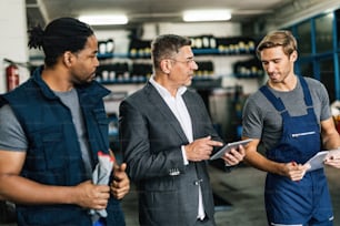 Hombre de negocios adulto medio que usa el panel táctil mientras habla con mecánicos de automóviles en un taller.