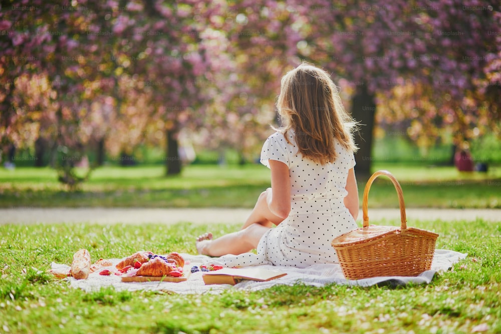 Beautiful young woman having picnic on sunny spring day in park during cherry blossom season