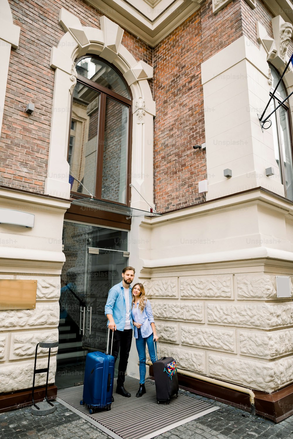 Picture of young couple entering hotel. Young man and woman standing near the old building city hotel, outdoors, holding suitcases.