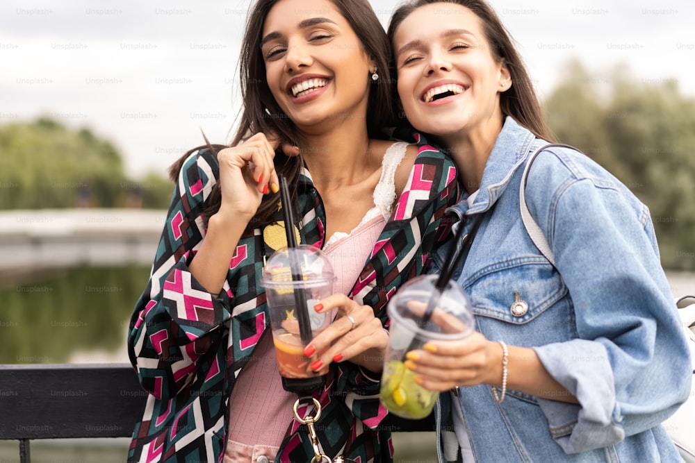 Two trendy cool hipster girls, friends drink cocktail in urban city background