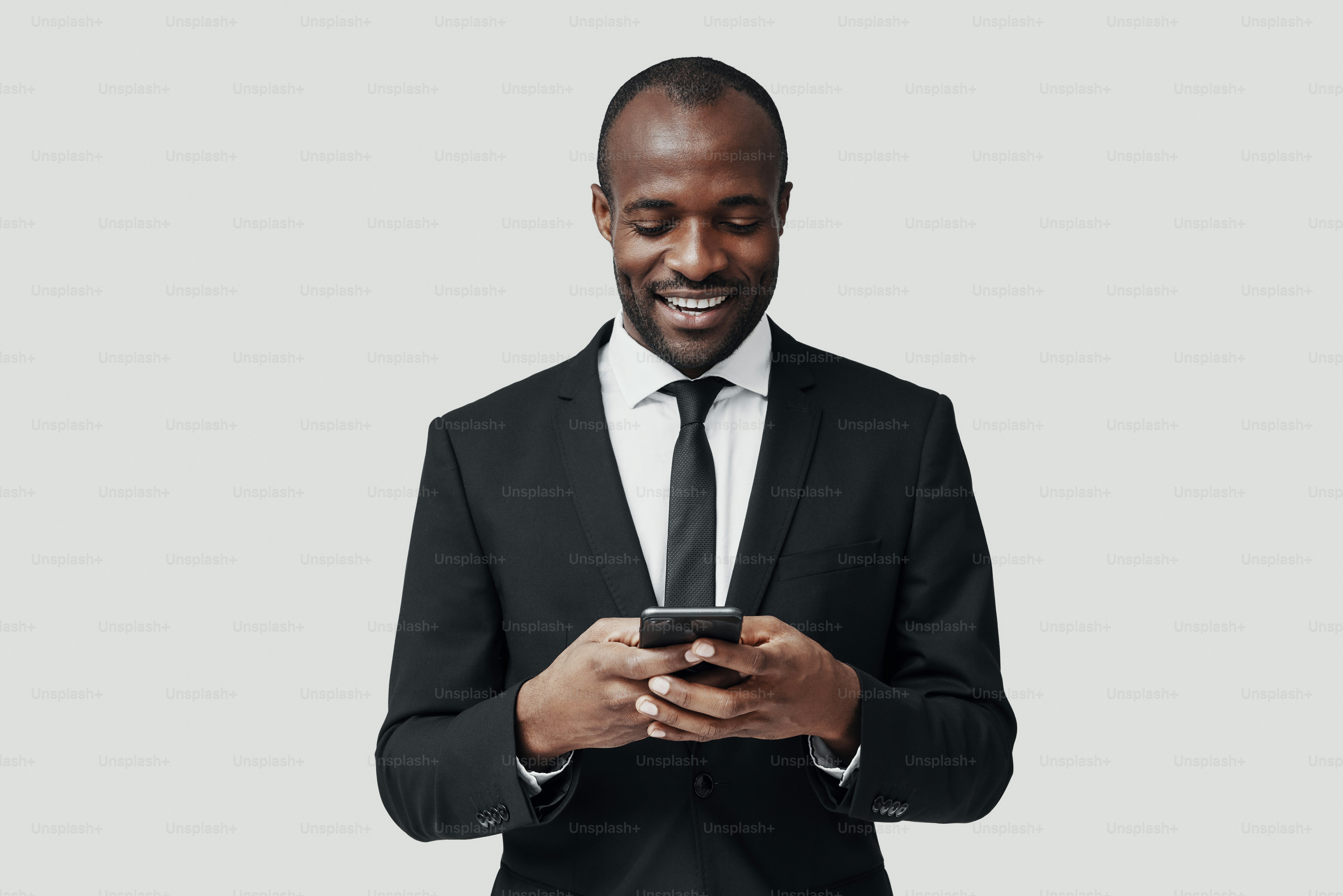 Modern young African man in formalwear using smart phone and smiling while standing against grey background